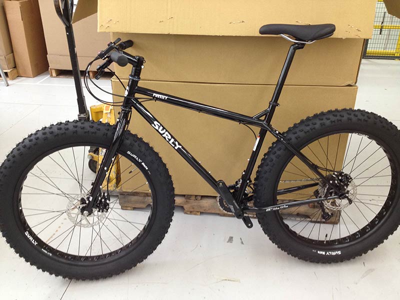 Left side view of a black Surly Pugsley fat bike with offset forks, leaning on a pallet of boxes in a warehouse