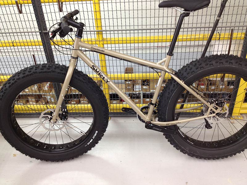 Left side view of a tan Surly Moonlander fat bike with offset forks, on a concrete floor, against a wire cage wall