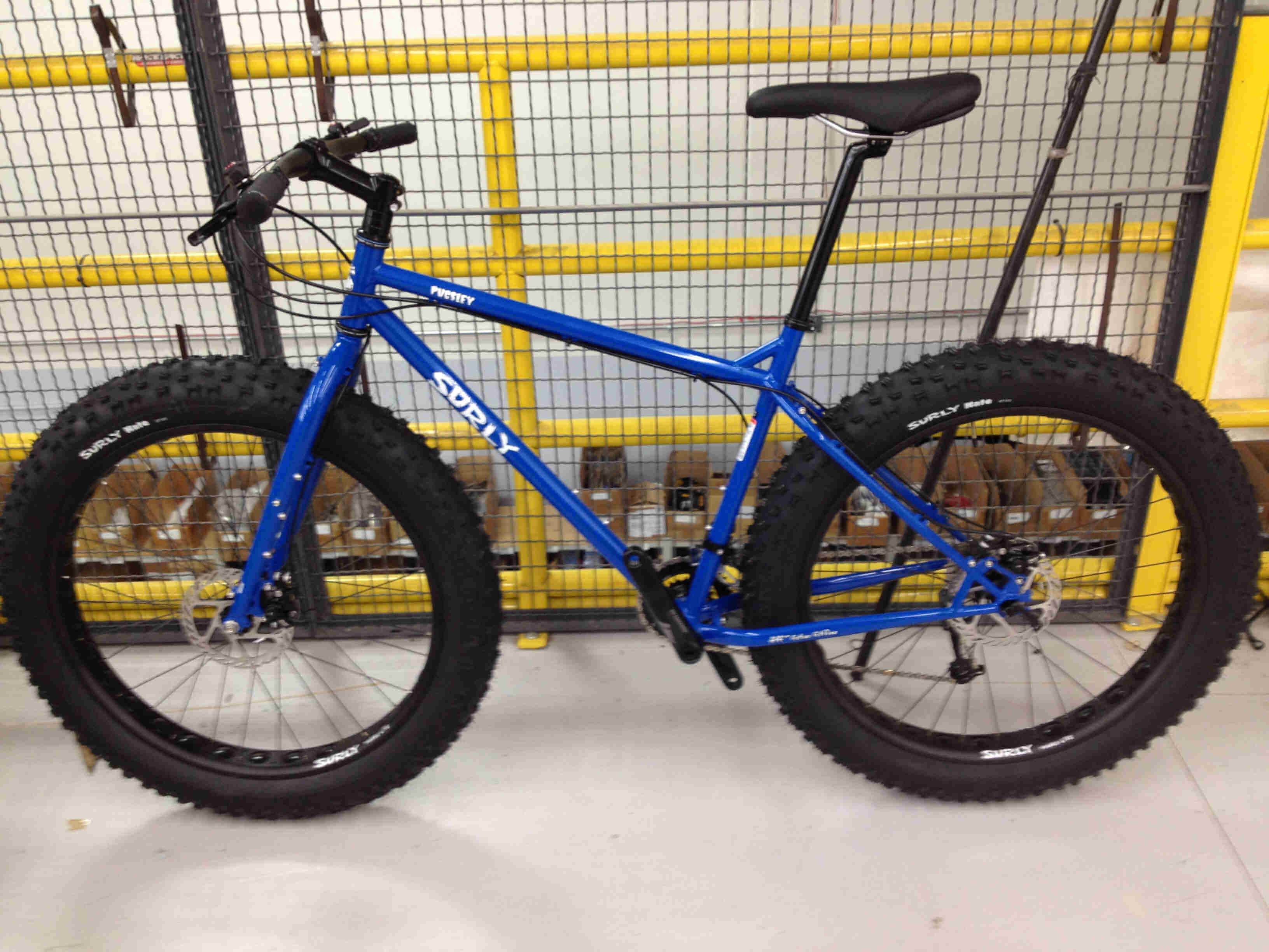 Left side view of a blue Surly Pugsley fat bike, on a concrete warehouse floor, against a wire cage wall