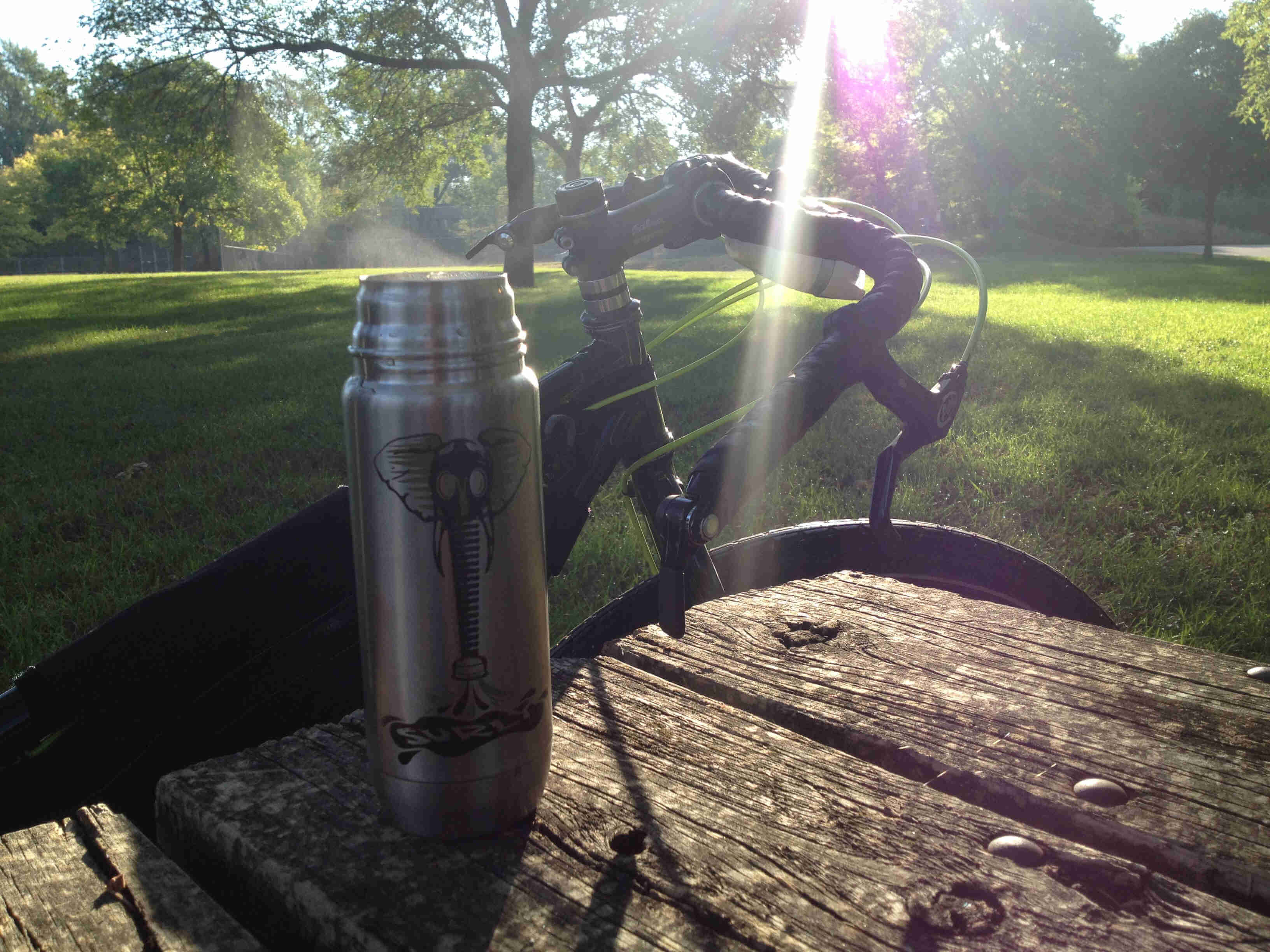 A steel Surly bikes water bottle with an elephant graphic on it, on a wood table with a bike behind it, in a park field