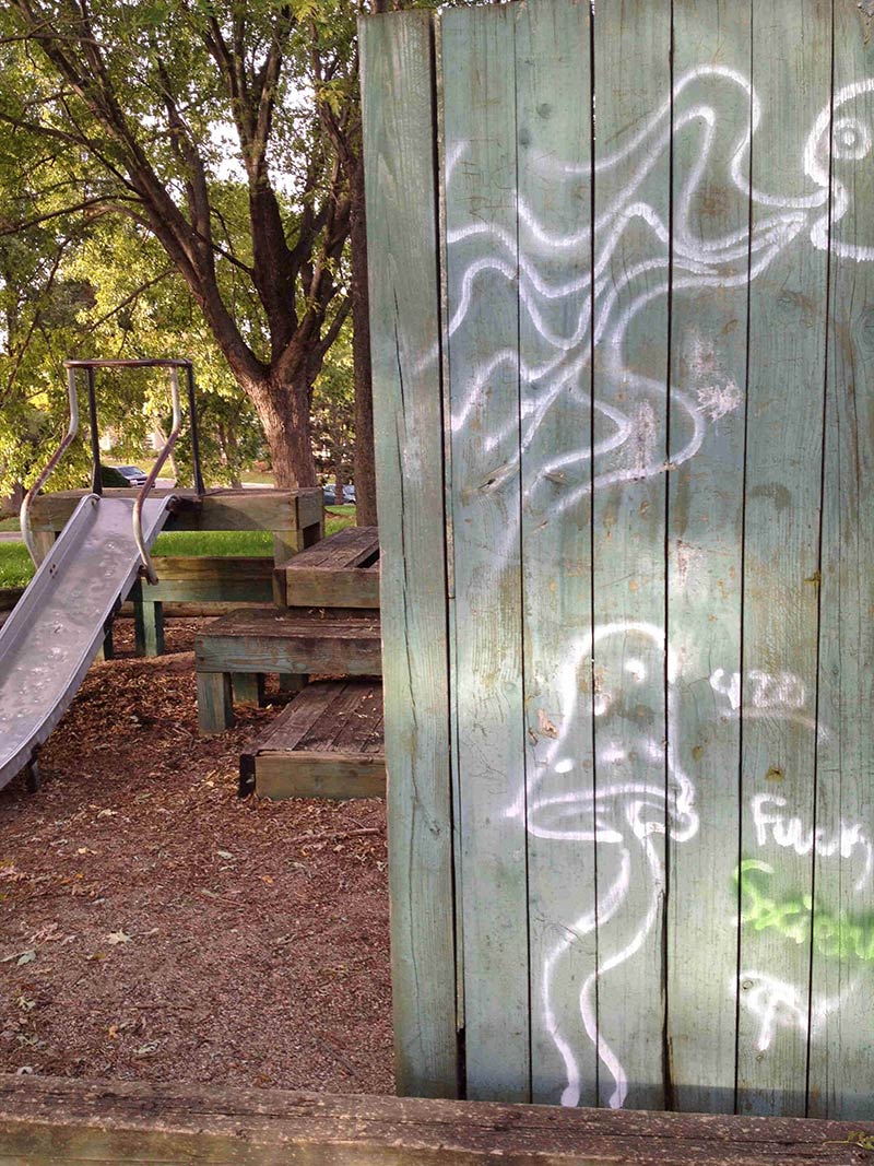 A wood fence wall with white spray painted graffiti on it, next to a wood playground structure with a slide on it