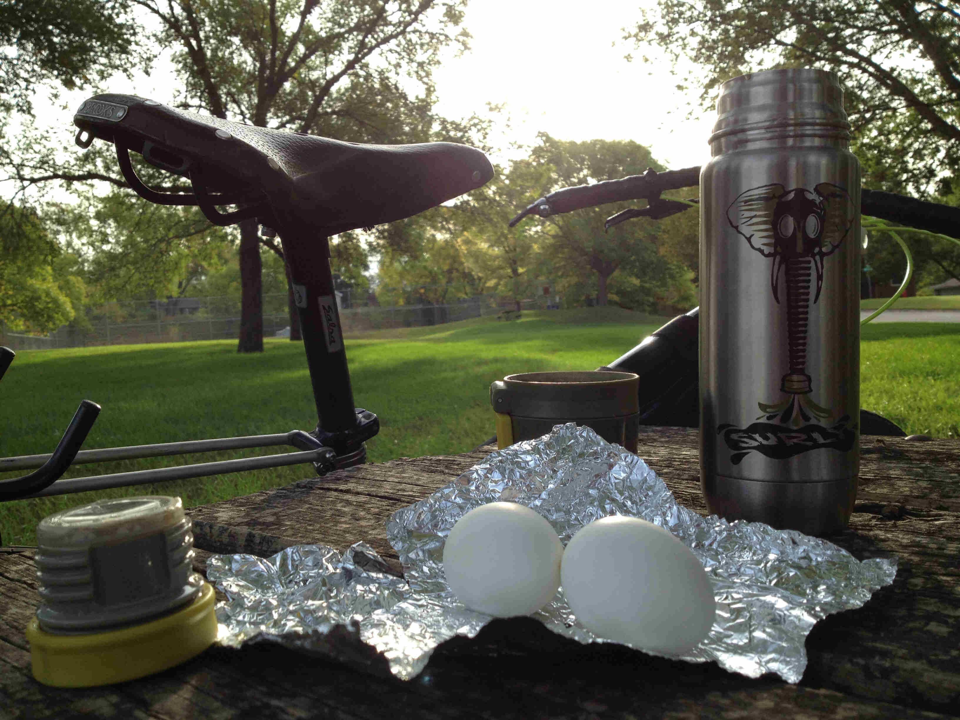 A wood picnic table with water bottle components and boiled eggs on foil, on the top, with a bike behind, in a park