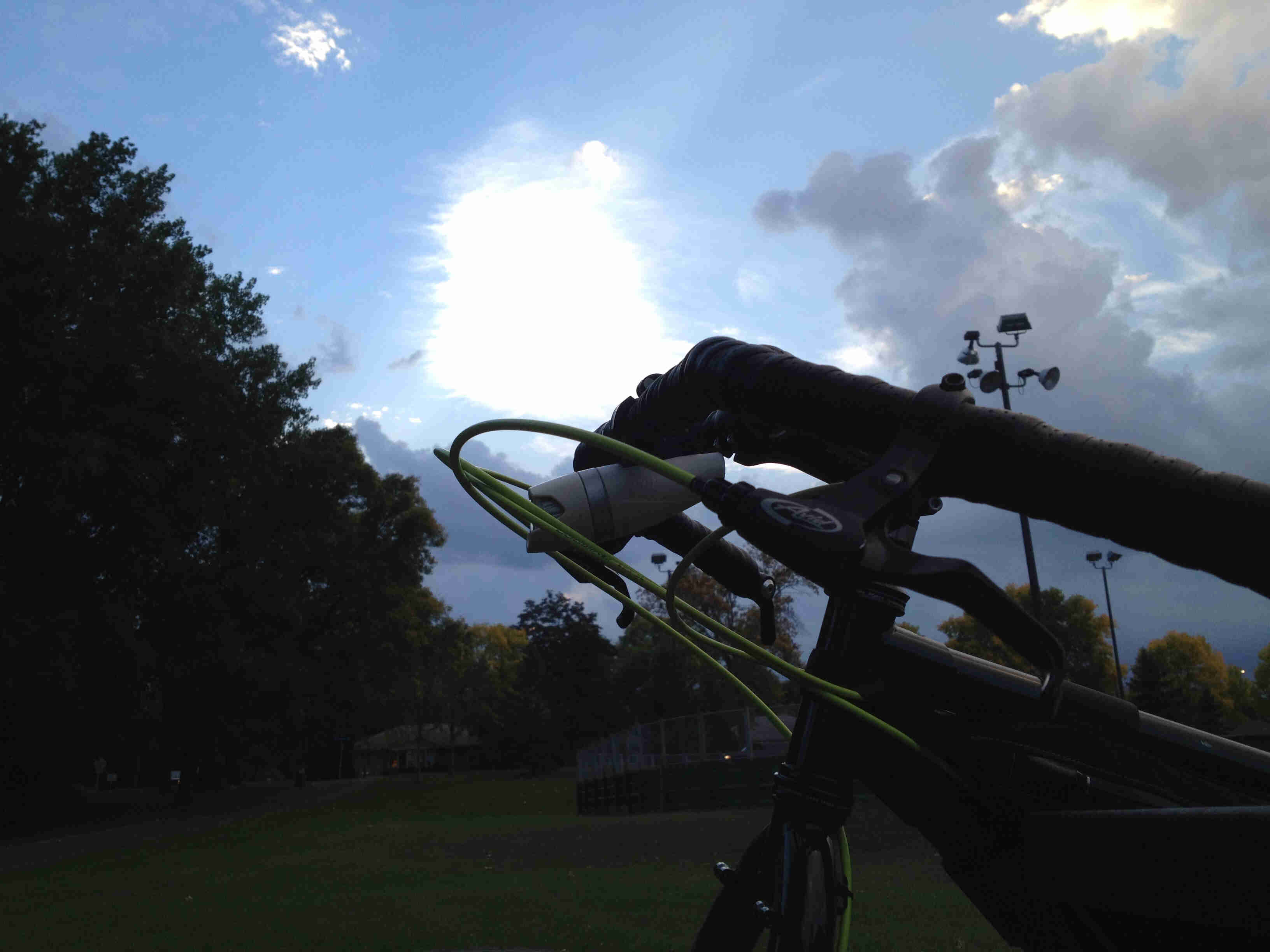 A dimly lit view of the upper, front part of a Surly bike, parked in a grass playing field with trees in the background