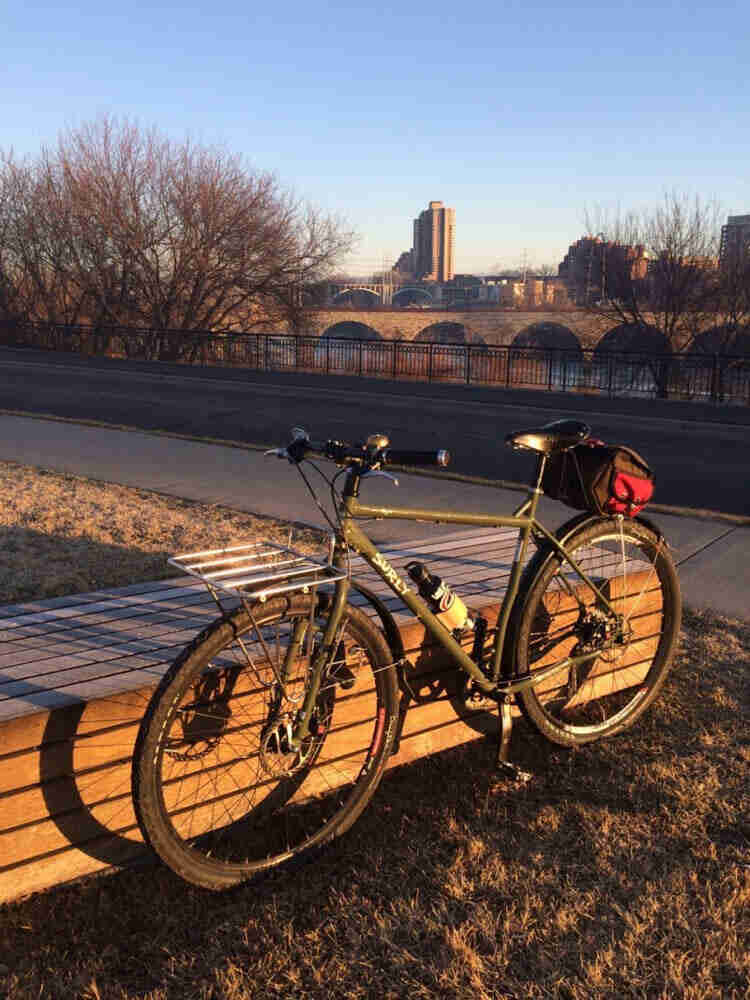 Left side view of a Surly bike, parked on grass in front of a bench, with a sidewalk, bridge and city in the background