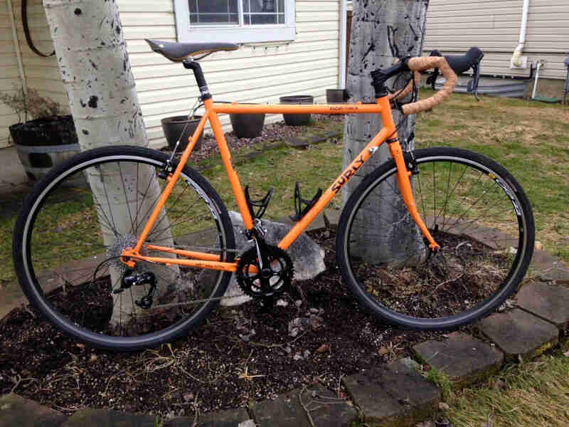 Right side view of an orange Surly Cross Check bike, parked in front of 2 trees in a background