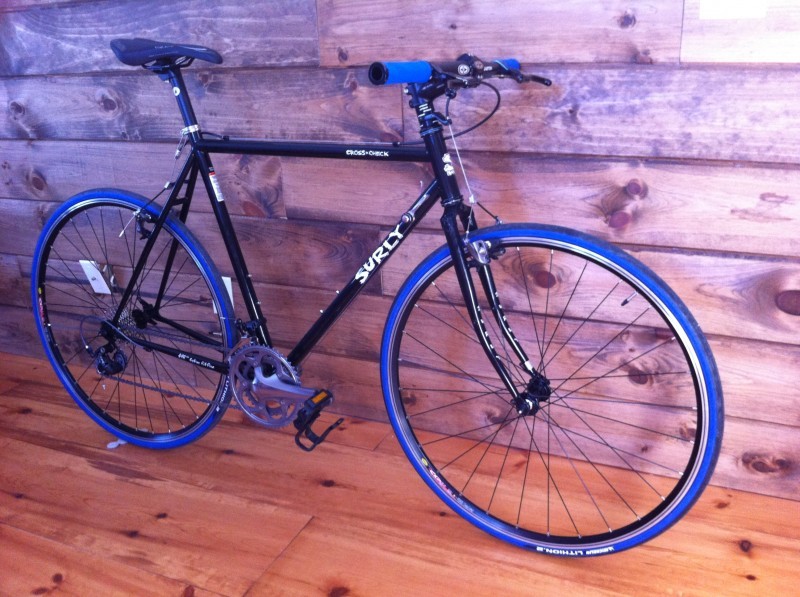 Right side view of a black Surly Cross Check bike with blue walled tires, leaning against a wood sided wall