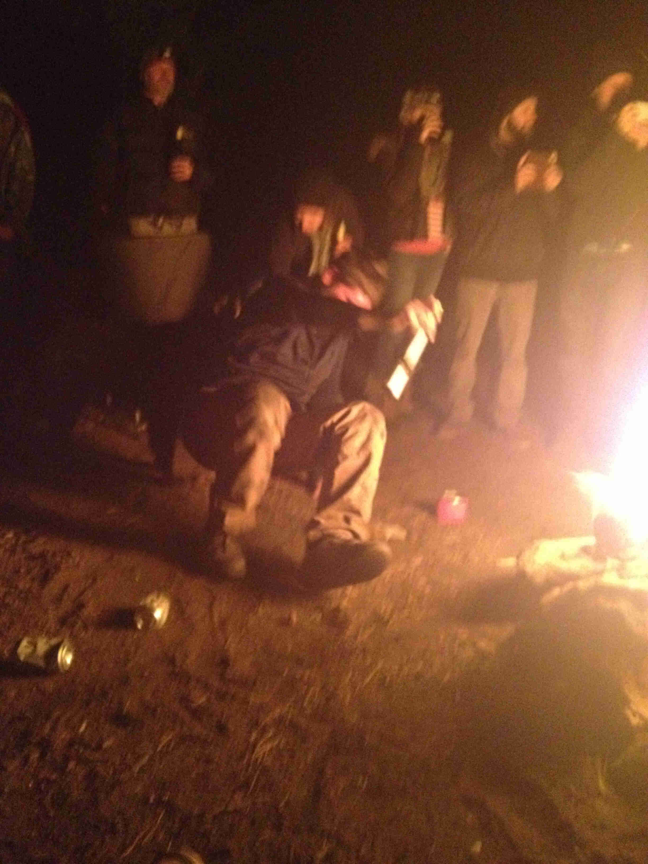 Front view of a person on the ground break dancing, next to campfire at night, with people standing behind them