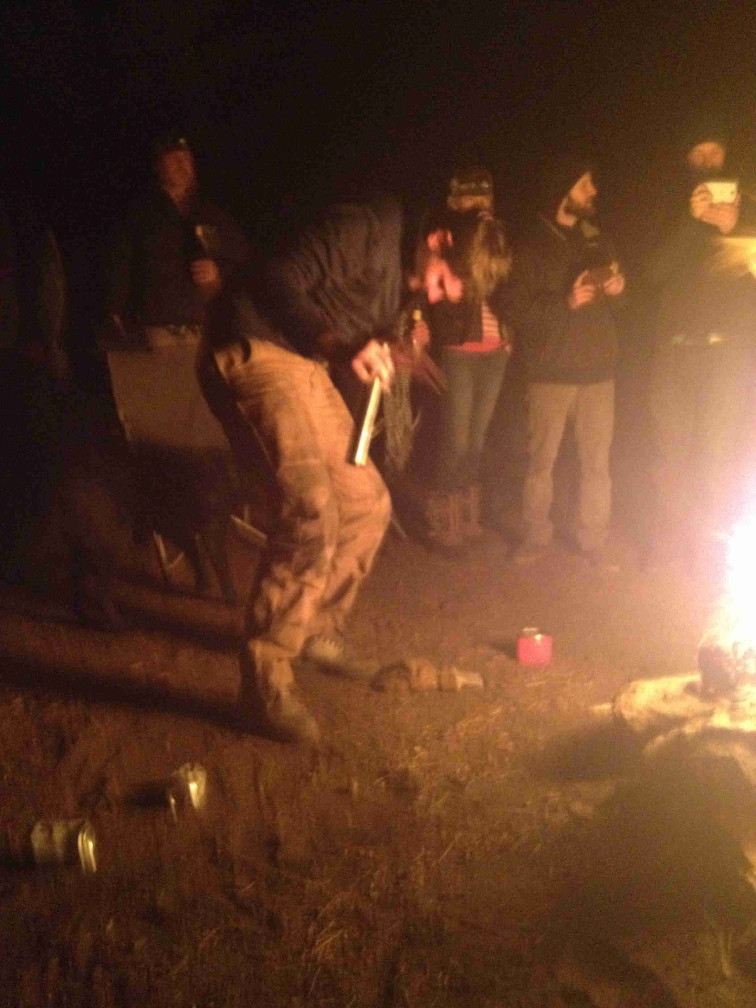 A person dancing next to a campfire at night, with others standing around in the background