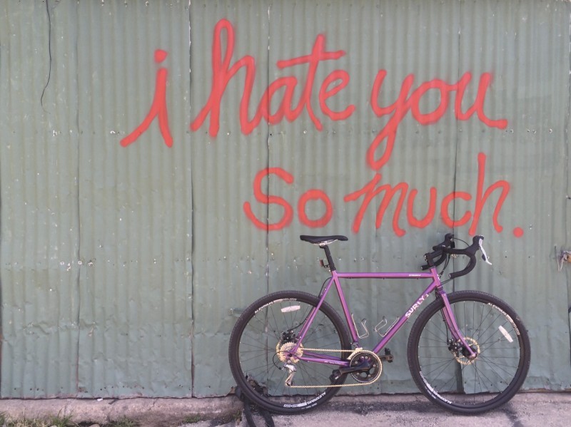 Right side view of a purple Surly bike, leaning on a steel panel wall with, I hate you so much, spray painted on it 