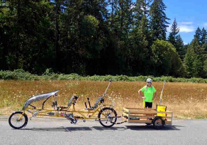 A cyclist standing behind a trailer attached to a parked recumbent bike, with a field and trees in the background