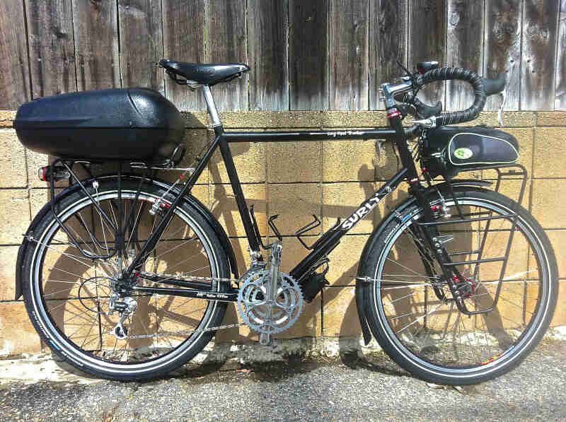 Right side view of a black Surly Long Haul Trucker bike with a rear hard case, parked against of block and wood wall