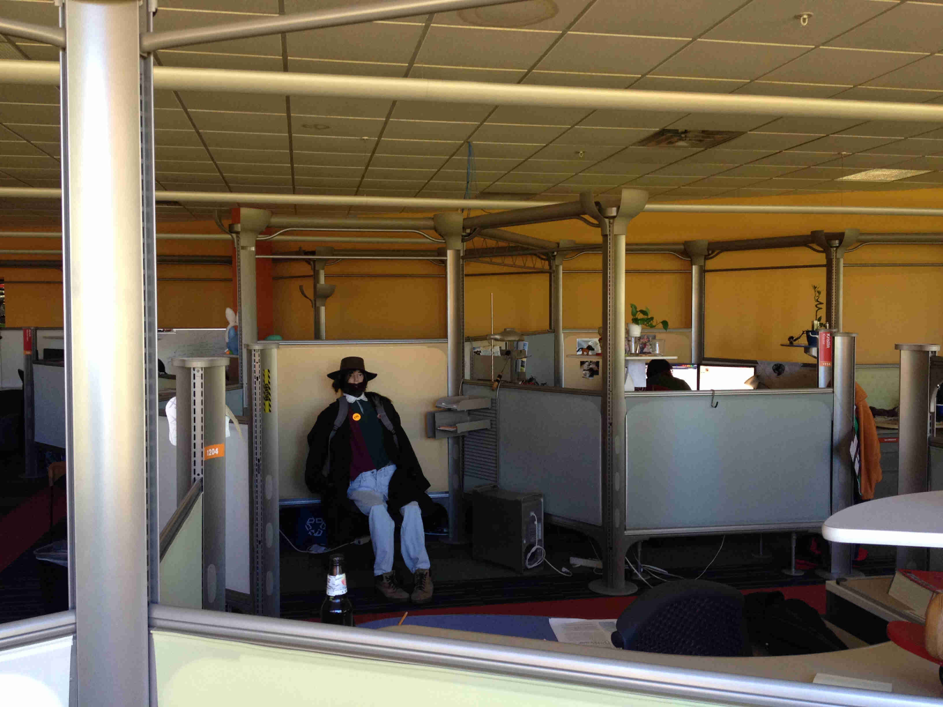 An unlit office room with cubicles, with a dressed up mannequin sitting on a bench, outside one of the office cubes