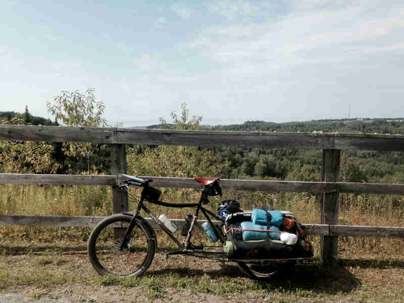 Left side view of a Surly bike with the back racks loaded with gear, parked in front of a wood rail fence