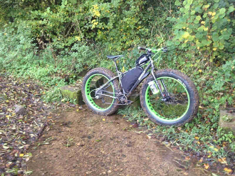 Right side view of a silver Surly fat bike with a frame bag, parked at the base of a cliff that's covered in weeds