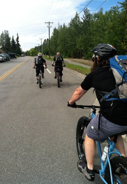 Rear view of a cyclist, riding a blue Surly fat bike on a street, with other riders in front and trees to the right