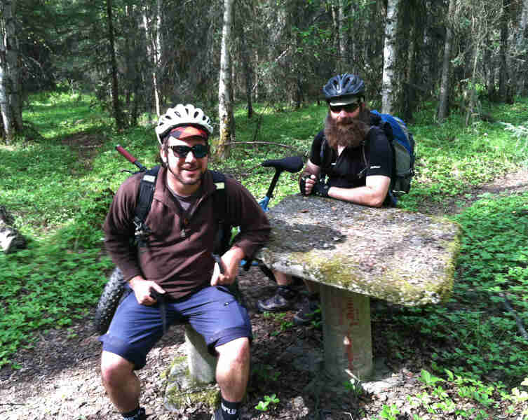 Front view of two cyclists sitting at a concrete table, with a Surly fat bike behind them, in a grassy, forest clearing