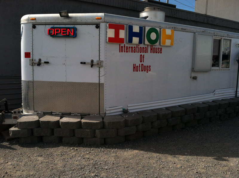 Side view of a white trailer on landscape block, with a neon OPEN sign, and International House of Hot Dogs on it