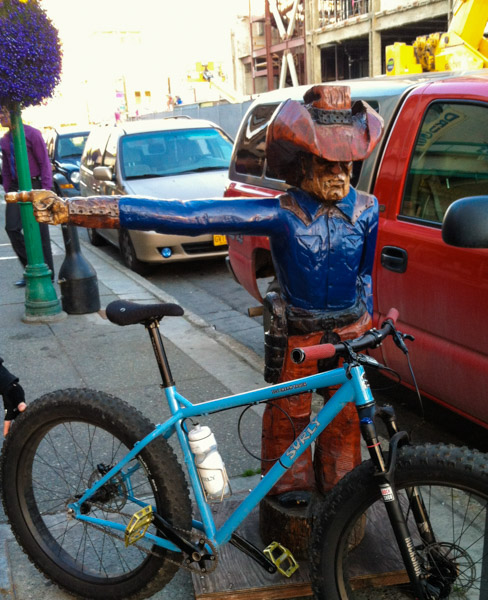 Right side view of a blue Surly Ice Cream Truck fat bike, leaning against a wooden cowboy statue, on a sidewalk