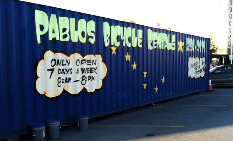 SIde view of a blue shipping container, on a parking lot, with PABLOS BICYCLE RENTALS, painted in mint lettering