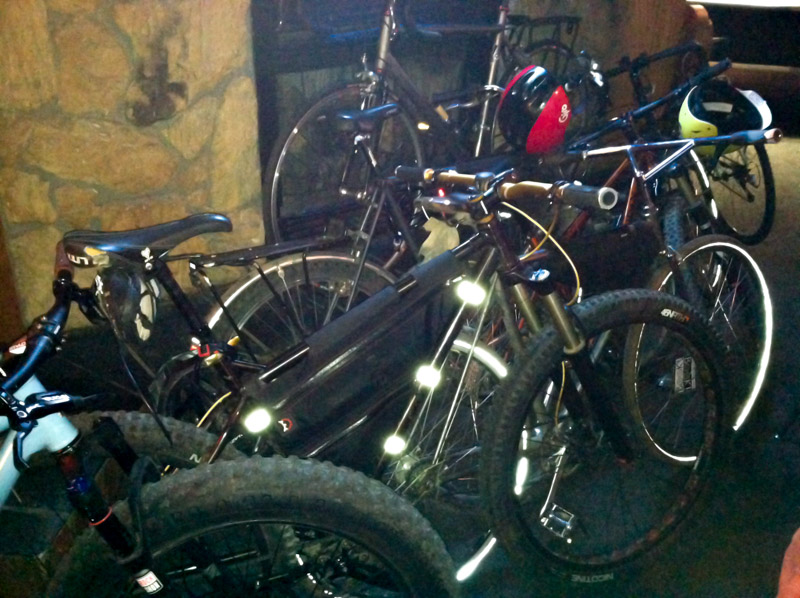 A collection of bikes, parked parked side by side, next to a stone wall at night