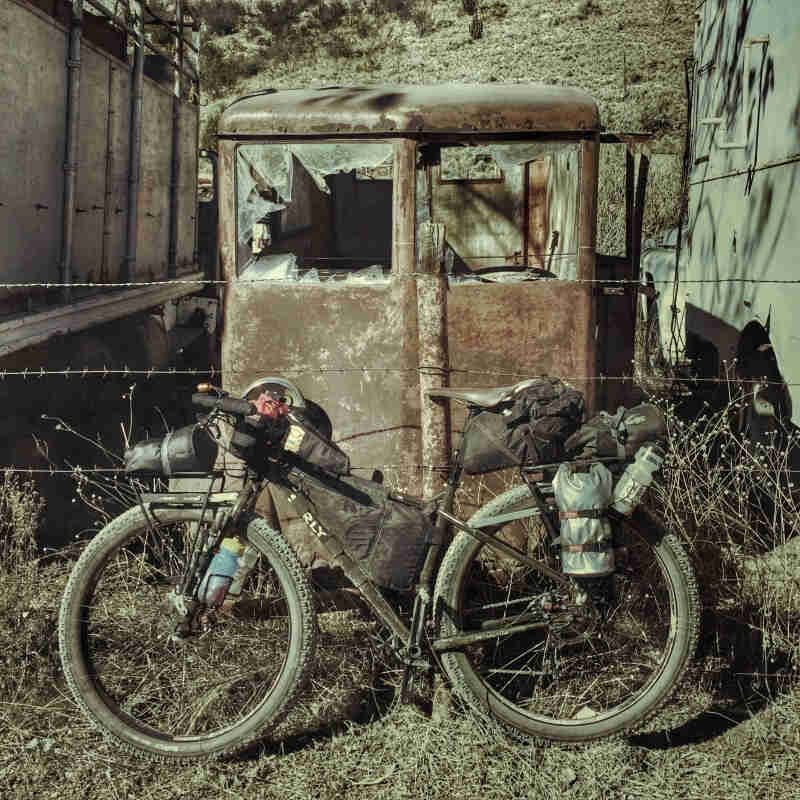 Left side view of a Surly bike with gear, in front of a wire fence with a rusty old truck with broken windshield, behind