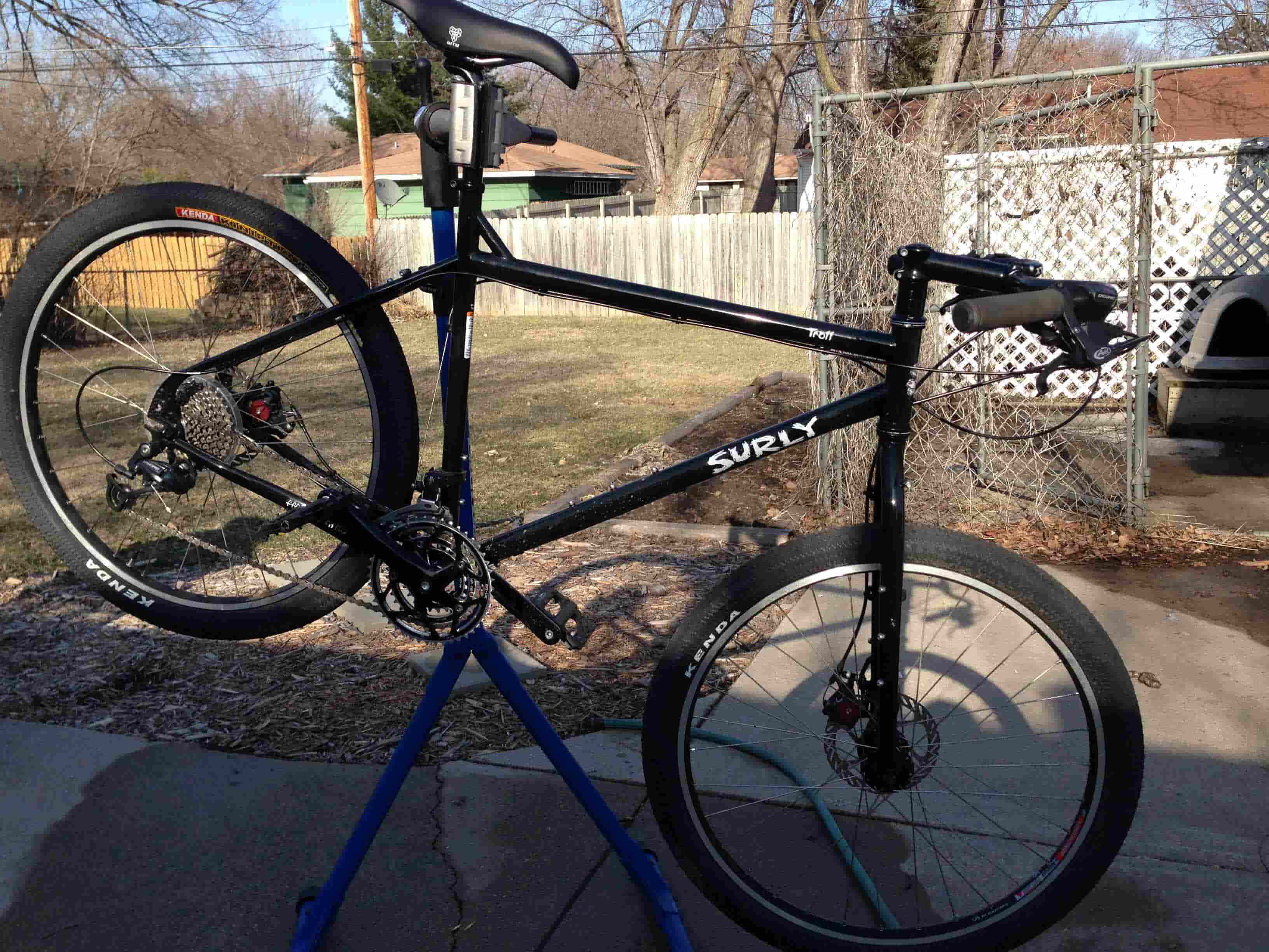 Right side view of a black Surly Troll bike, on a bike repair stand, parked on concrete beside a yard with a wood fence