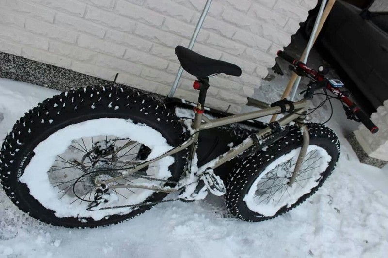Right side view of a tan Surly Moonlander fat bike, with snow on the rims, leaning against an exterior brick wall