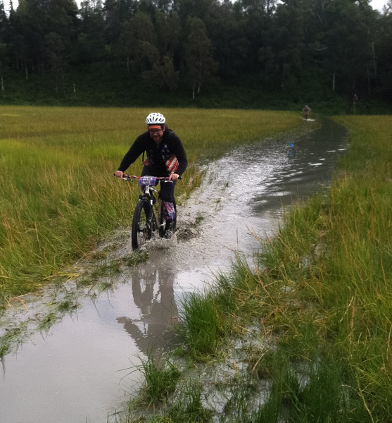 Front view of cyclist riding through a narrow waterway, in a wetlands field, with trees in the background