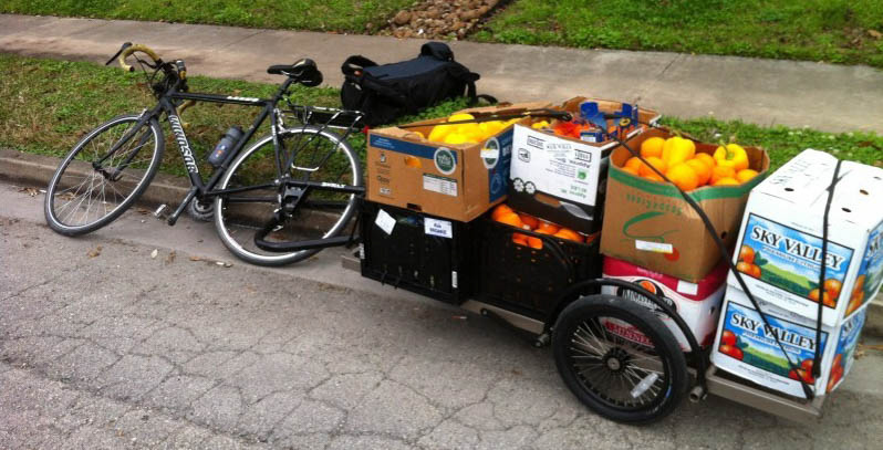 Left side view of a bike laying on it's side, with a trailer of vegetables attached, parked on the side of a street