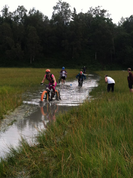 Front view of cyclists riding through a narrow waterway, in a wetlands field, with trees in the background