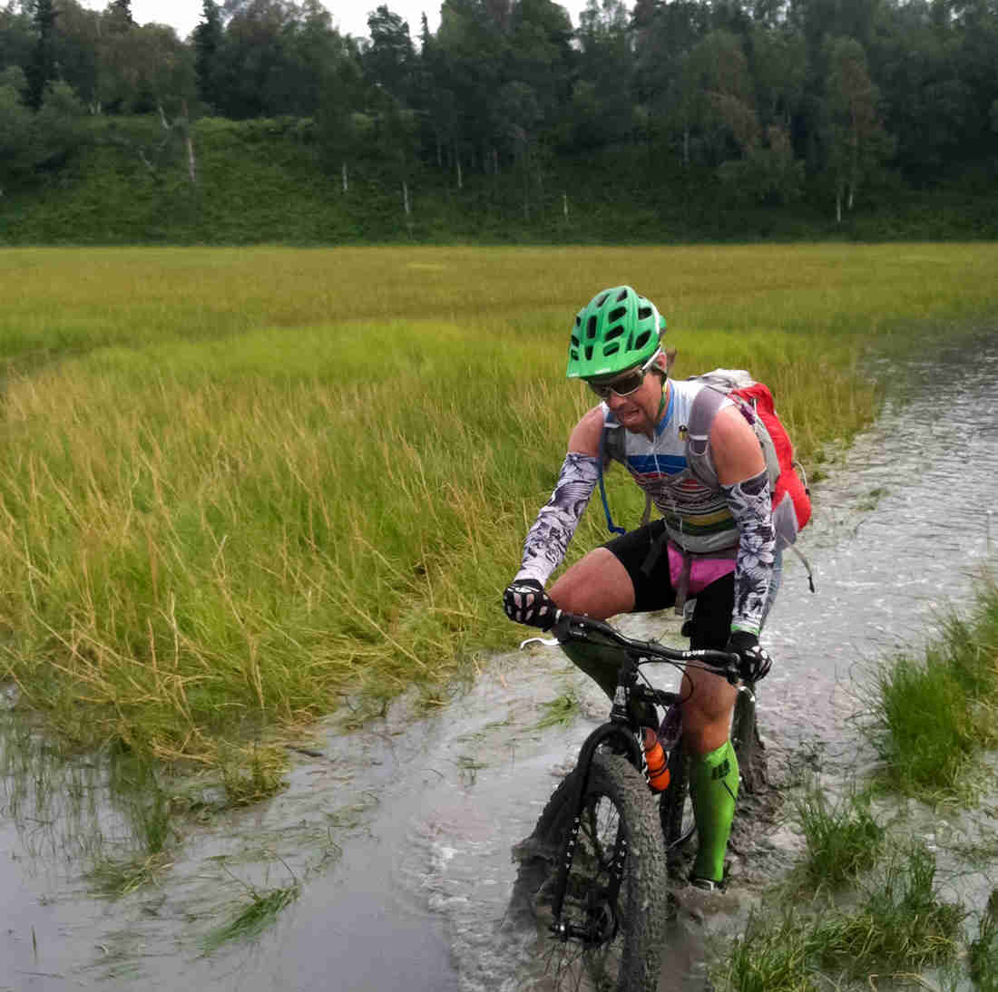 Front view of a cyclist, riding a black Surly fat bike through a waterway in a wetlands field, with trees in background