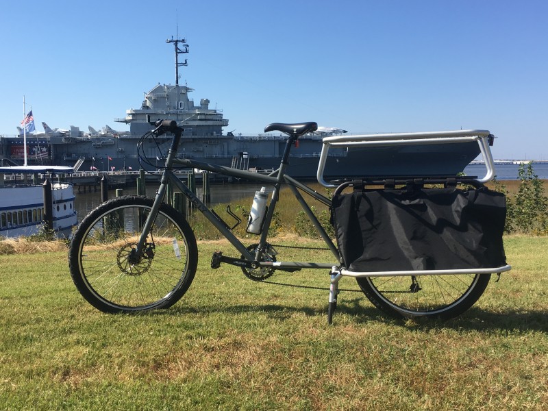 Left side view of a Surly Big Dummy bike in a grass field, with a aircraft carrier in the background