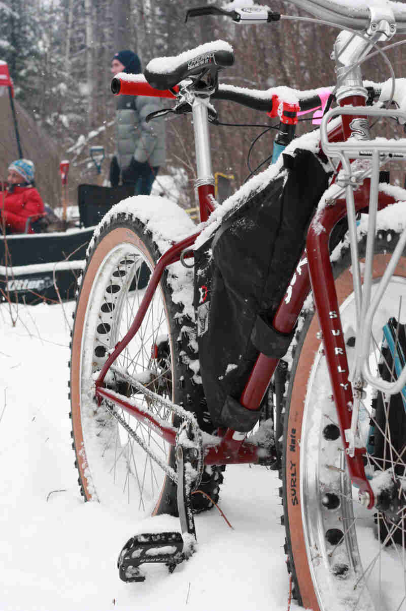 Close up, front right side view of a red Surly fat bike standing in snow, with people and trees in the background