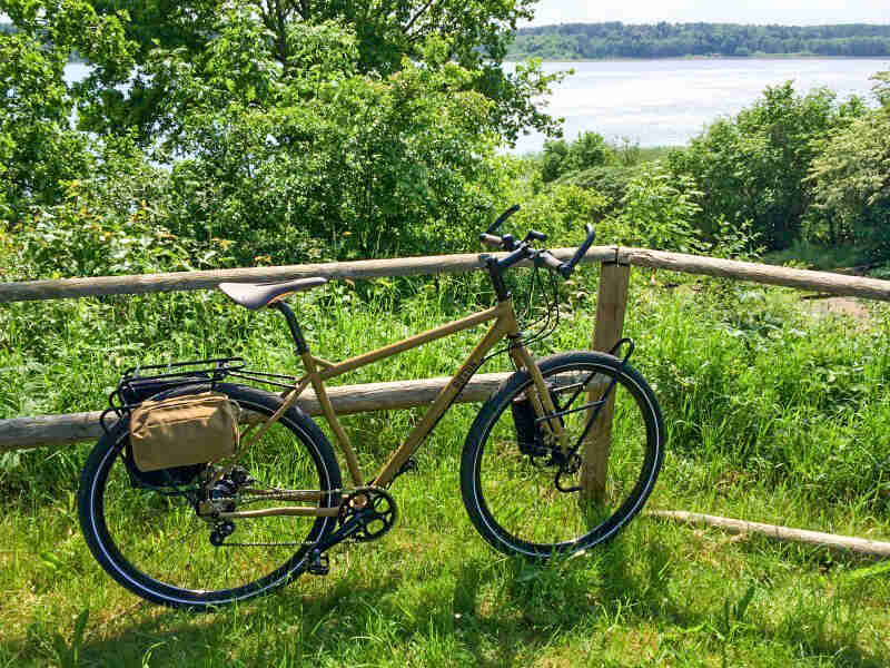 Right side view of a brownish yellow Surly bike, parked along a log fence, with thick trees and a lake in the background