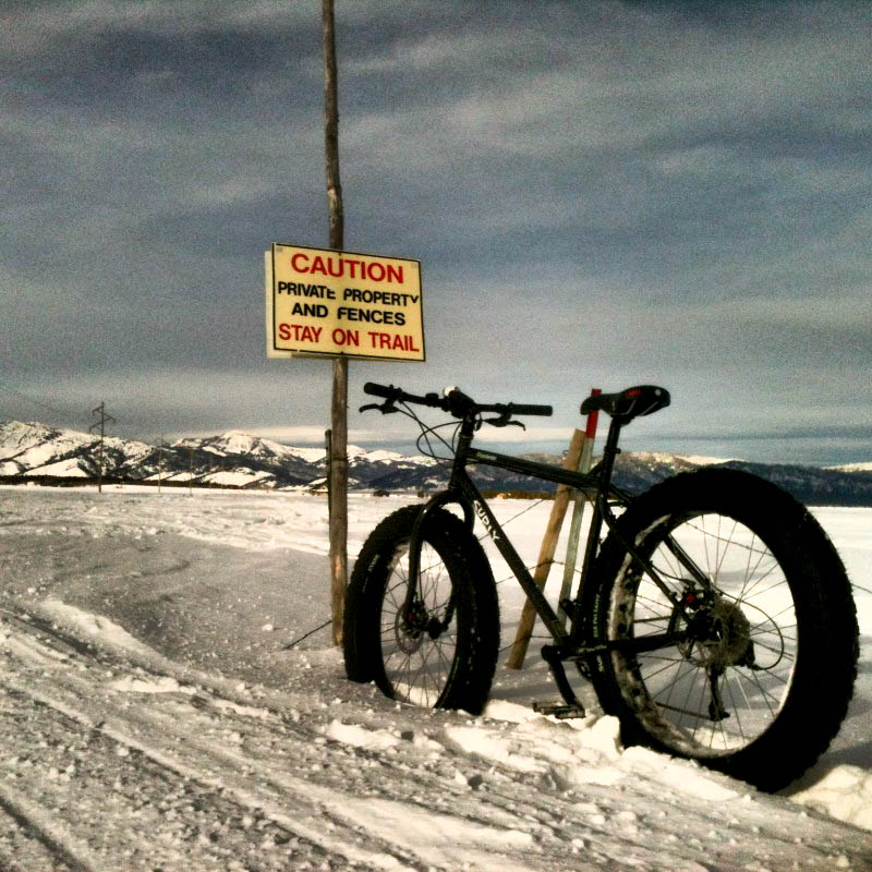 Left side view of a Surly fat bike, parked in a field of deep snow, in front of a sign, with mountain in background