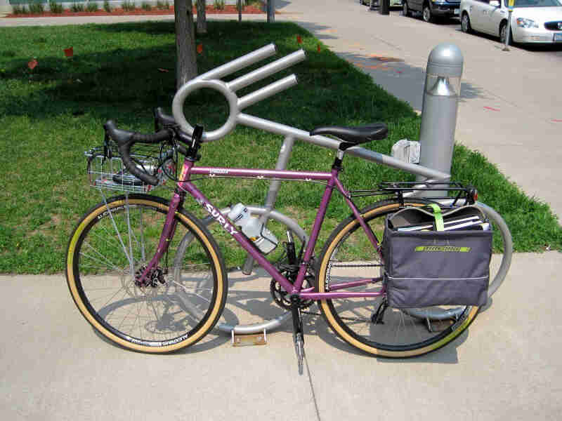 Left side view of raspberry color Surly bike with a rear pack, parked alongside a bike rack on a sidewalk
