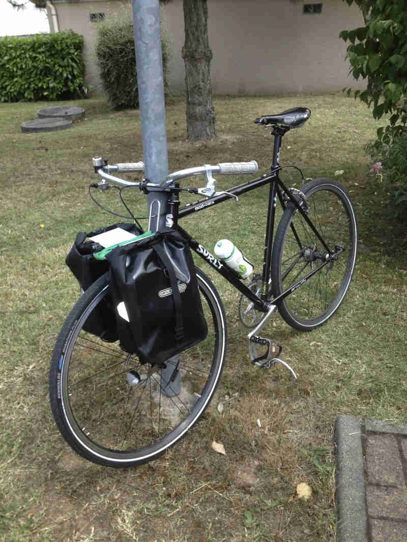 Front, left side view of a black Surly Cross Check bike with front saddlebags, leaning against a pole in a grass area