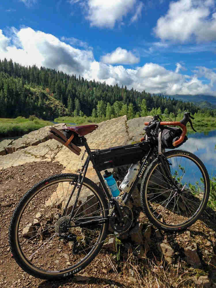 Right side view of a black Surly bike, in front of a slanted rock, with a lake and a pine forest in the background