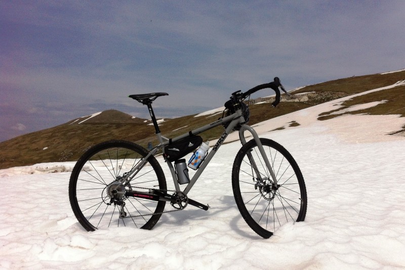 Right side view of a gray Surly Karate Monkey bike, parked on a large patch of snow, on the hill of a mountain