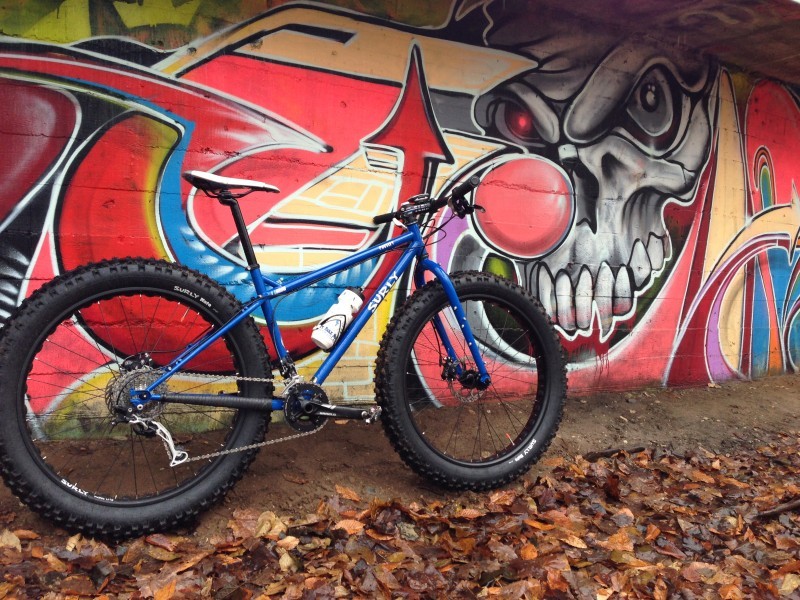 Right side view of a blue Surly fat bike, in front of a cinder block wall with mural painted on it