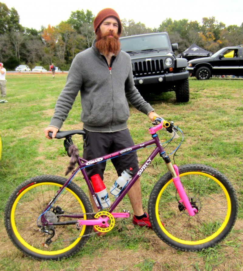 Right side view of a purple Surly Troll bike with yellow rims, and a cyclist standing behind, in a grass field
