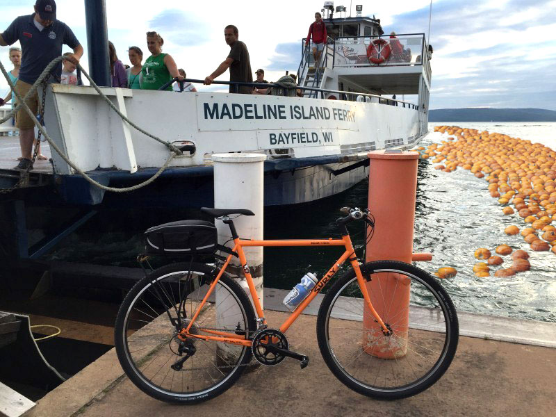 Right side view an orange Surly Cross Check bike, on a dock next to a ferry boat with people - pumpkin graphics in water