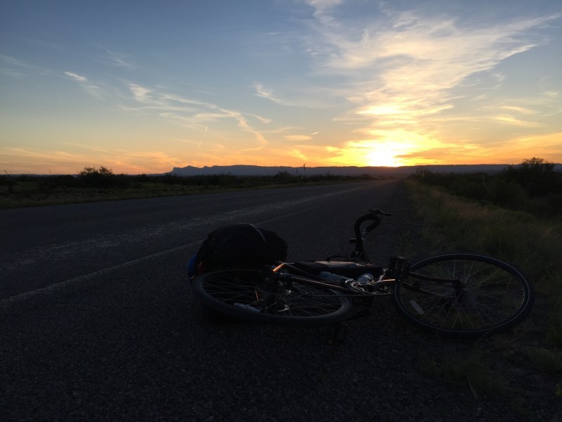 A bike, loaded with gear, laying on it's left side on a roadside, with the sun on the horizon in the background