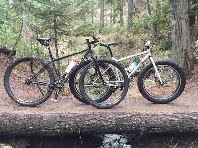 Right side view of a black Surly bike, with a white Surly fat bike behind it, parked on a log trail bridge in the forest
