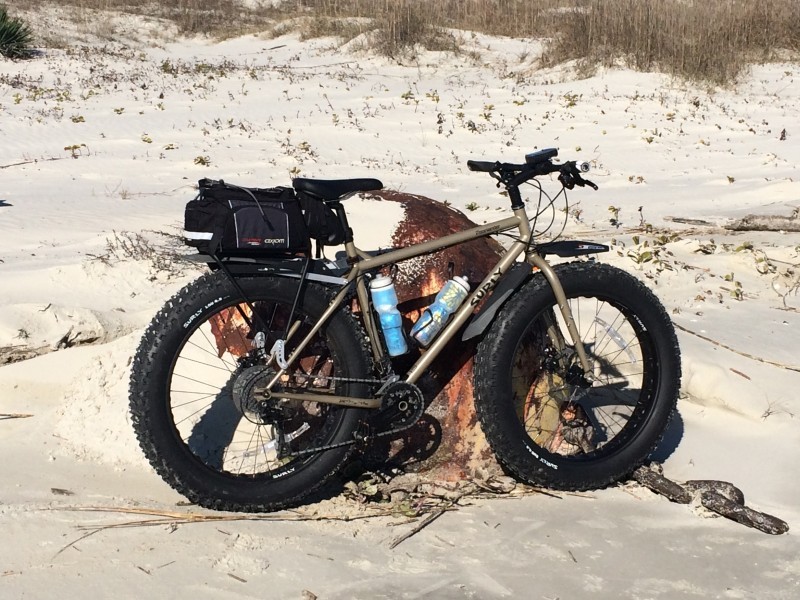 Right side view of a tan Surly Moonlander fat bike, parked against a large, rusty steel ball, on a beach