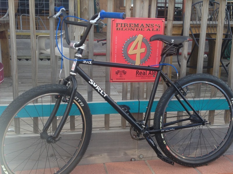 Left side view of a black Surly 1x1 bike, leaning against balusters with a sign on them, on the outside of a wood deck