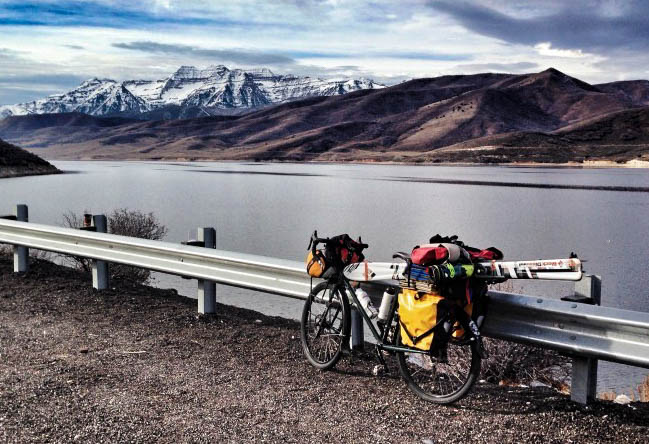 Left side view of a bike loaded with gear and skis, parked along a guardrail, with a lake and mountains in background