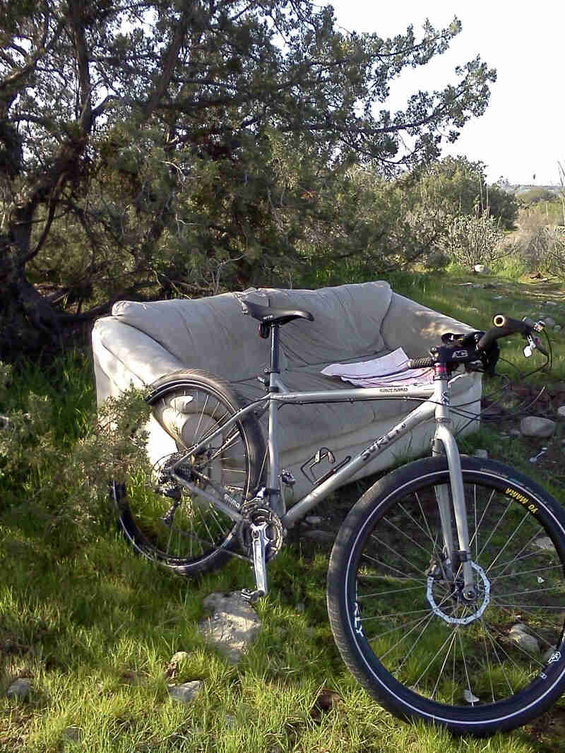 Right side view of a gray Surly Karate Monkey bike, leaning on a couch in a rocky, grass clearing, with a tree behind it