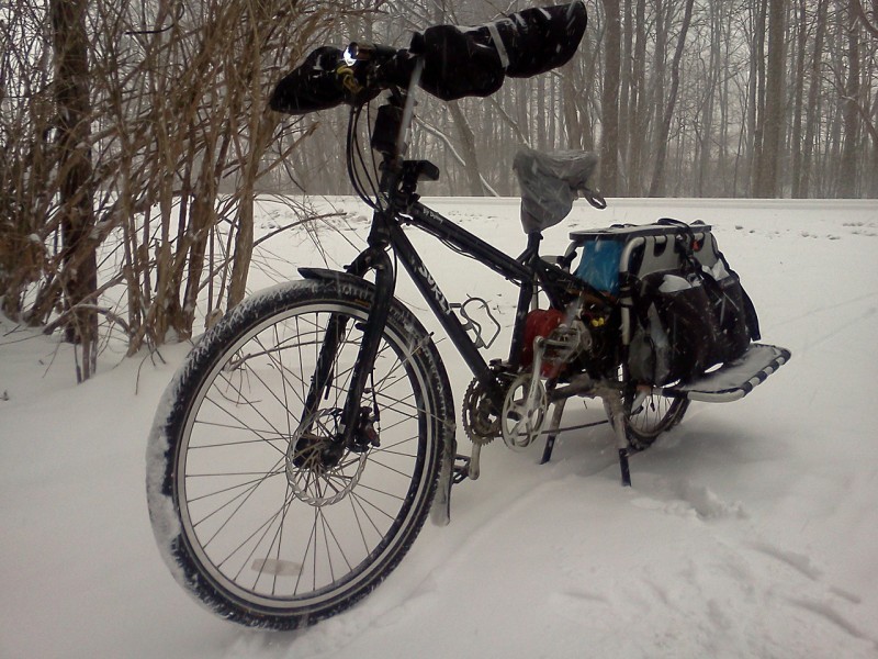 Front, left side view of a black Surly Big Dummy bike, standing in deep snow, with bare trees in the background
