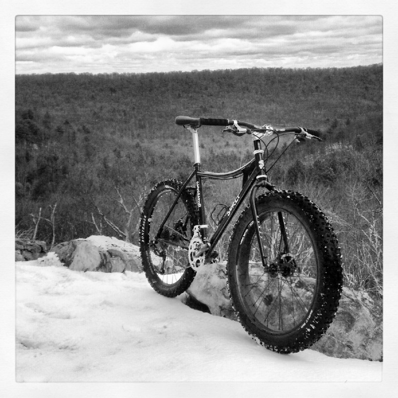 Front, right side view of a Surly Instigator bike, on a snowy hilltop ledge, overlooking a forest below on a cloudy day